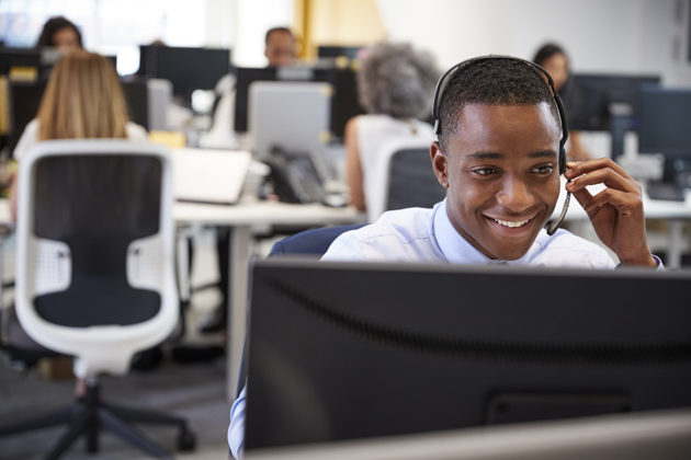 A smiling man wearing a headset is working at a computer in a modern, open-plan office.